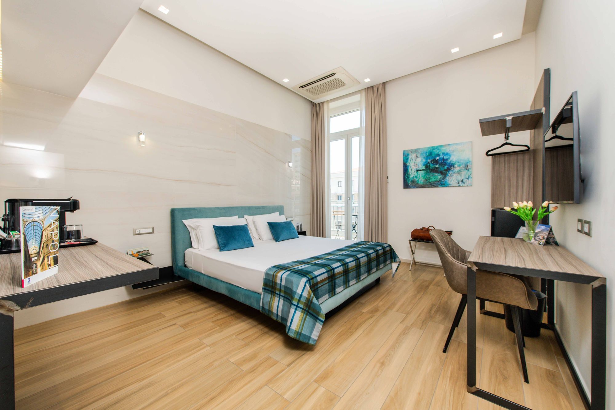 Letto matrimoniale con plaid a quadri e testiera turchese, scrivania con TV a parete, macchina di caffè
