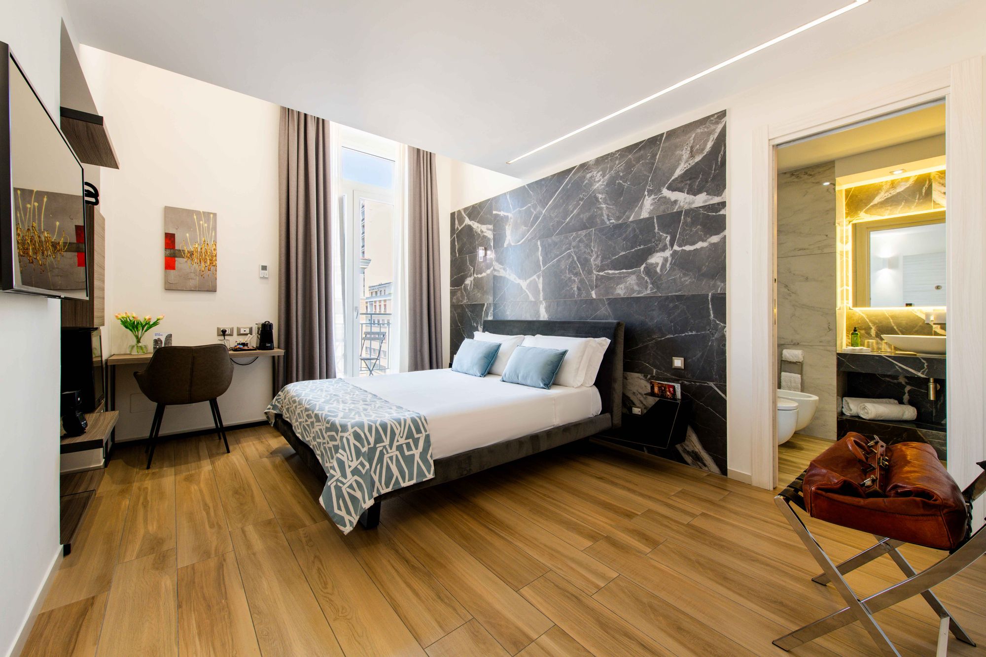 Camera con parete in marmo, letto con cuscini azzurri e copriletto geometrico, scrivania vicino alla finestra - Municipio Luxury Suite Napoli, foto 1